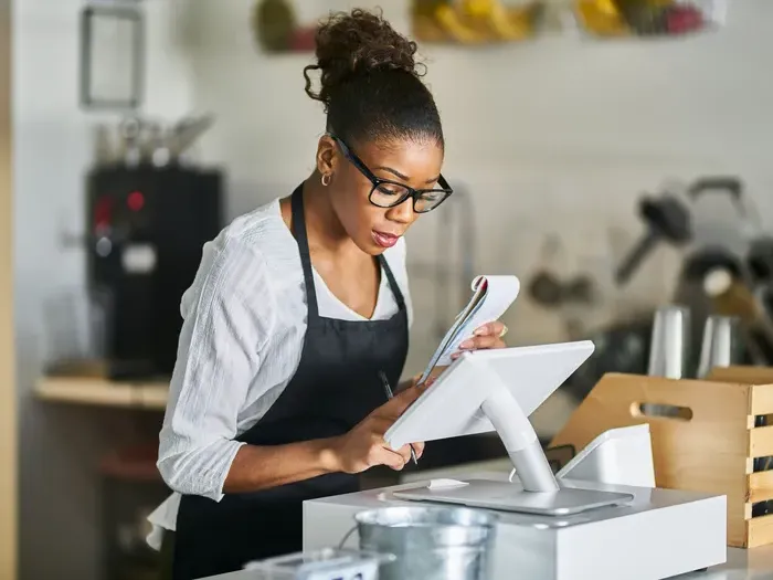 Shop owner checks orders on a touchscreen POS at the counter, representing small business tools and listings